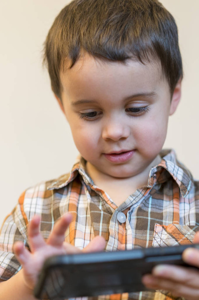 Portrét usměvavý chlapeček držení mobilního telefonu přes světlé pozadí, samostatný. roztomilé dítě hraní her na smartphonu. vertikální fotografie. Detailní záběr. - Fotografie, Obrázek