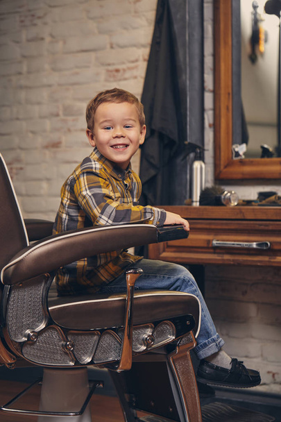 Retrato de um menino elegante vestido de camisa e jeans na barbearia, sentado em uma cadeira contra o local de trabalho barbeiros
 - Foto, Imagem