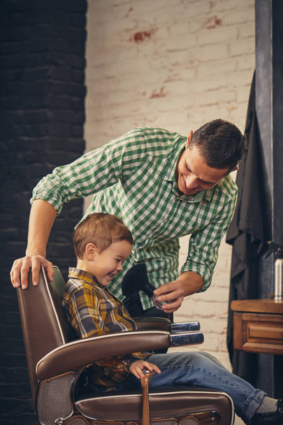stylish little kid sitting on chair at barbershop with his young father on background - Photo, Image