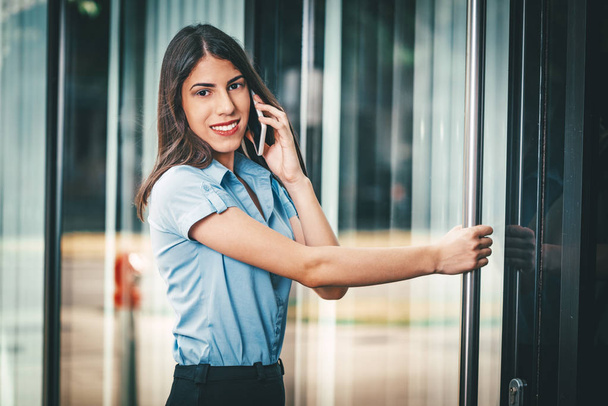 Joven mujer de negocios hablando en smartphone y de pie en la puerta del centro de negocios
 - Foto, Imagen