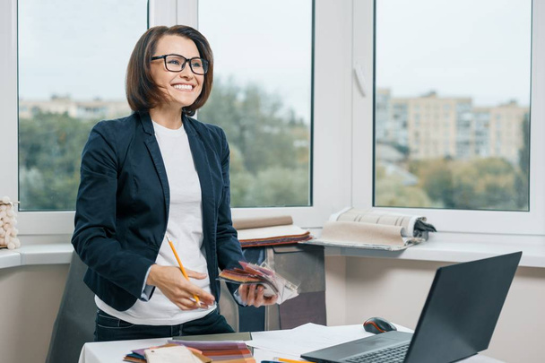 Nainen sisustussuunnittelija työpaikalla kangasnäytteitä, kannettava tietokone. Hymyilevä nainen takissa, lasit katsovat sivulle seistessään lähellä pöytää
 - Valokuva, kuva