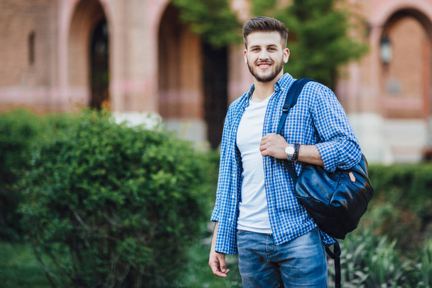 Joven hombre guapo en camisa azul con mochila de pie en el campus y mirando a la cámara
 - Foto, Imagen