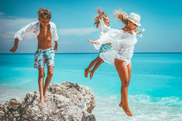 Happy young mother with little kids having fun at sunny beach while  jumping from rock  - Photo, Image