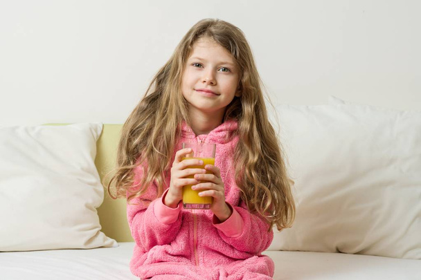 Girl child in pajamas with a glass of fresh orange juice sitting at home in bed - Zdjęcie, obraz