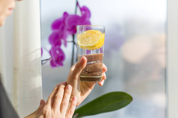 Mature woman is holding a glass with healthy drink. Natural antioxidant water with lemon, female stands near a window, enjoys drink - Foto, Imagem