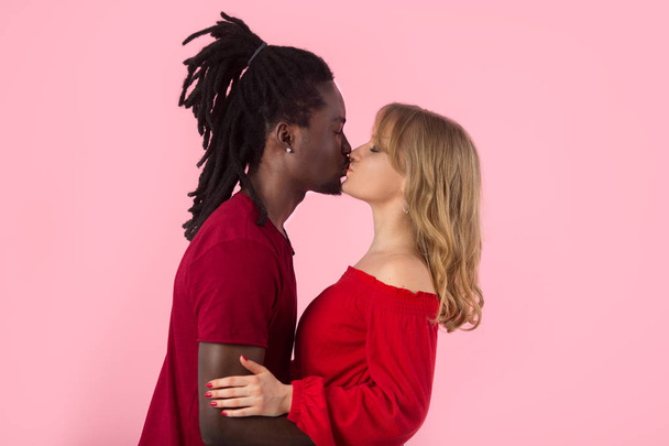 beautiful young couple african man and white woman in red t-shirts on pink background kiss - Photo, Image
