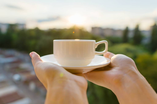 Female hands holding classic white cup with saucer, background evening sunset, city silhouette, golden hour - Фото, изображение