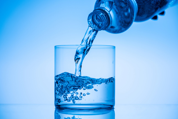 oříznutý pohled muž vylévající vodu z plastové láhve ve skle na modrém pozadí - Fotografie, Obrázek