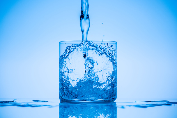 toned image of water pouring in drinking glass on blue background with splashes - Foto, afbeelding