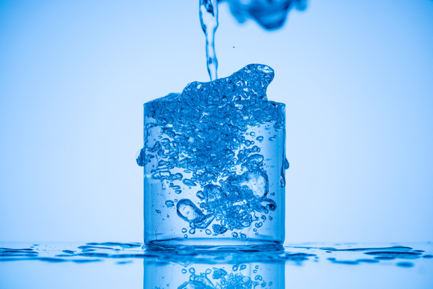 imagem tonificada de água derramando em vidro cheio sobre fundo azul com salpicos
 - Foto, Imagem