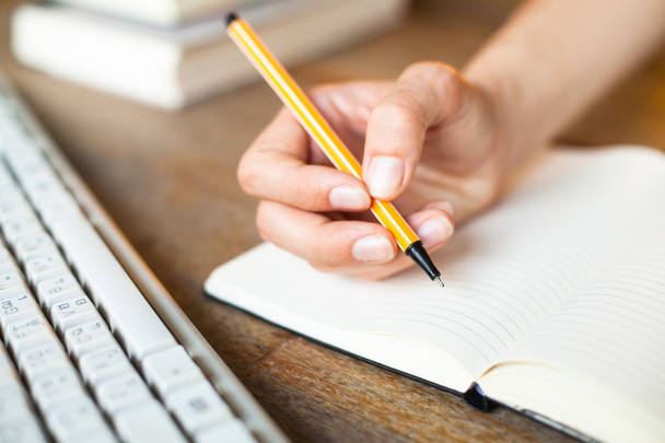 Hände schreiben einen Stift in ein Notizbuch, eine Computertastatur und einen Stapel Bücher - Foto, Bild