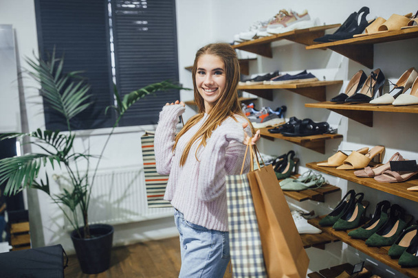 Young beautiful woman with shopping bags posing at rack with shoes in stylish store - Valokuva, kuva
