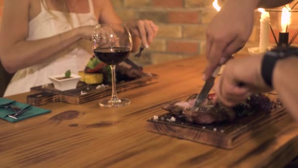 Man and woman cutting meat steak while dinner with candles in evening restaurant. Romantic couple dinning in barbeque restaurant. - Imágenes, Vídeo