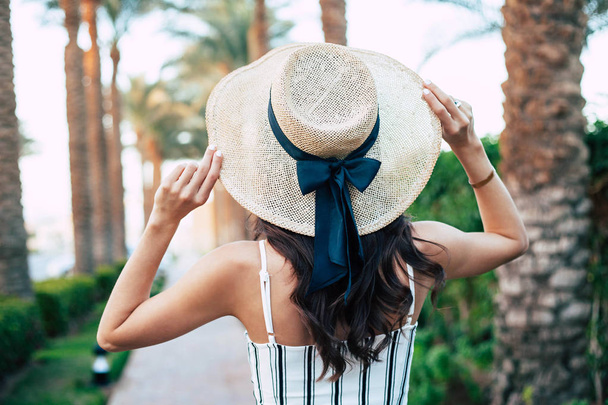 Facing the joy. Tanned girl among the beautiful palms and greenish bushes in camel hat decorated with a black bow and matching jumpsuit.  - Foto, afbeelding