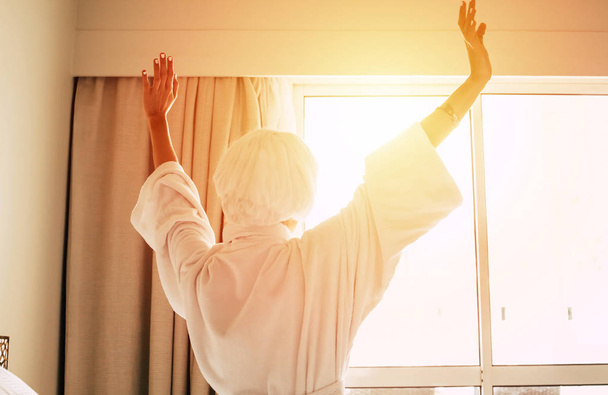 Εγρήγορσης. Υπέροχη γυναίκα σηκώνοντας τα χέρια της έως την ημέρα ηλιόλουστα κάθεται σε ευάερα φως δωμάτιο με μια πετσέτα μπάνιου και μπουρνούζι της. - Φωτογραφία, εικόνα