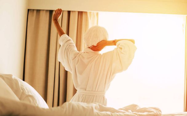 Magic of the morning. Magnificent woman is pulling her hands up to get some of the power of the sunlight and sitting on the bed with a light bed clothes on it. - Foto, Imagen