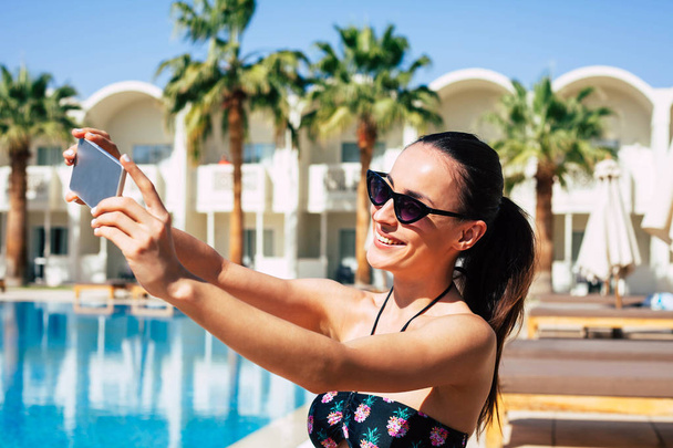 Follow me! Hilariously beautiful girl is taking a selfie next to the swimming pool ,greenish palms,white building and wooden lounge chaises. - Фото, изображение