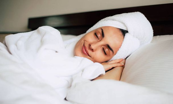 Middle day nap. Cute young girl is having a relaxing nap after shower in cozy double bed with bright white bedclothes. Her skin is tanned, eyebrows are nicely shaped and her smile is pleasant. - Valokuva, kuva