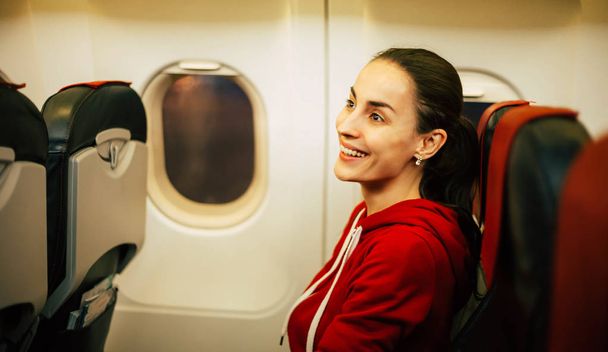 Finally here. Side-drawn photo of fabulous girl with stunning smile and sincerely happy eyes sitting in the airplane and wearing a comfortable ruby hoodie. - Foto, immagini
