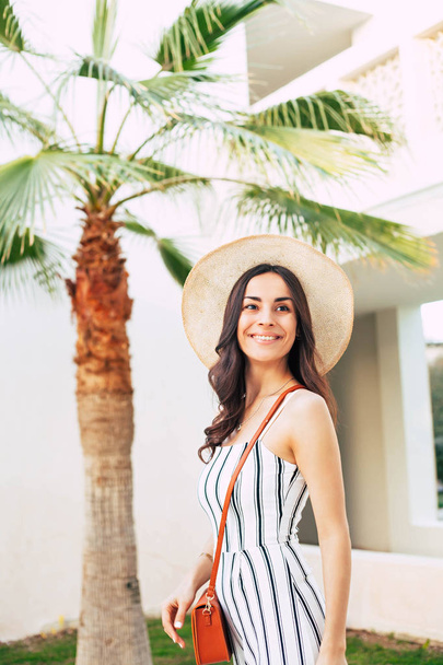 Slender girl near the green palm tree in a beige hat and striped jumpsuit with a foxy coloured bag over her shoulder. She has a genuinely smile on her face and her hazel eyes are full of bliss. - Valokuva, kuva