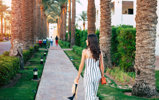 As I wish. Calm gorgeous girl walking through surrounding beauty of vibrant green palms and bushes in pretty black & white outfit and leather shoes with foxy coloured bag. - Фото, изображение