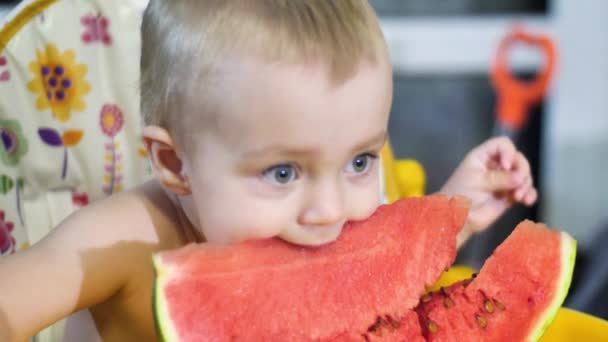 beautiful baby boy sitting at the kids table on the kitchen looking in the camera and then bites a piece of watermelon close up view slow mo video in 4K - Filmati, video