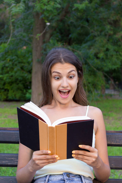 Mujer joven sorprendida con un amplio abierto sí y la boca está leyendo un libro al aire libre
 - Foto, Imagen