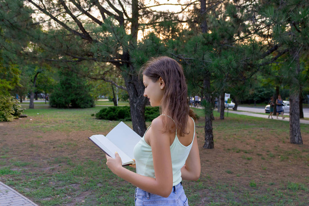 Kaunis naisopiskelija kävelee puistossa ja lukee kirjaa.
 - Valokuva, kuva