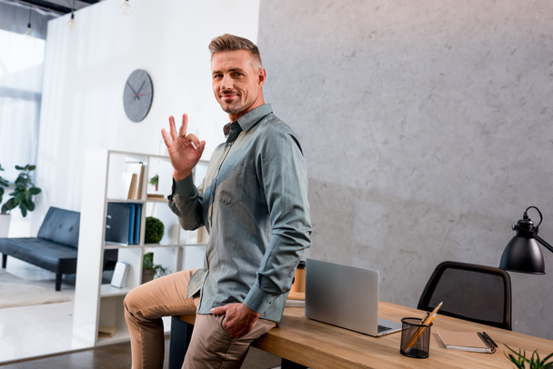 happy businessman showing ok sign while sitting with hand in pocket in office  - Foto, immagini