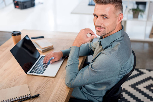 handsome businessman using laptop with blank screen while looking at camera - Photo, Image