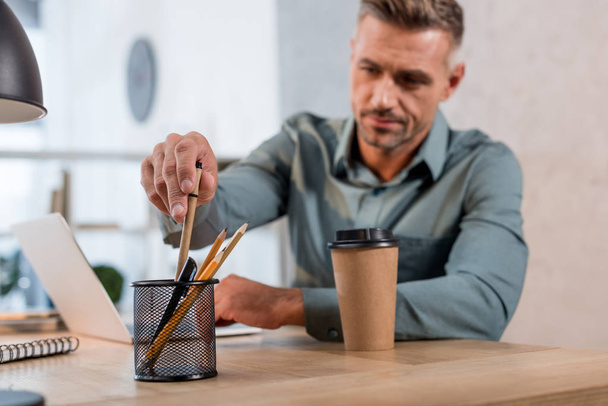 selective focus of man taking pen from metallic holder with stationery near paper cup in office - Fotó, kép