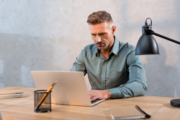 pohledný muž sedí u stolu a používat přenosný počítač v kanceláři - Fotografie, Obrázek