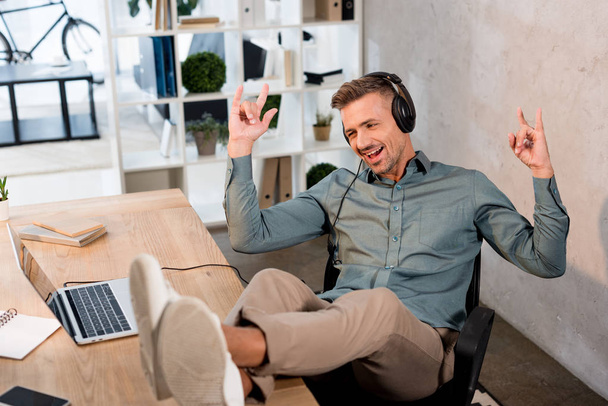 happy businessman listening music in headphones while showing rock sign in office - Photo, Image