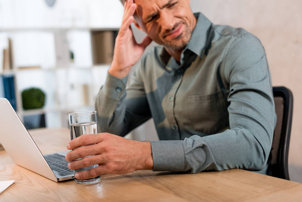 tired businessman holding glass of water while having headache in office - Foto, Imagem