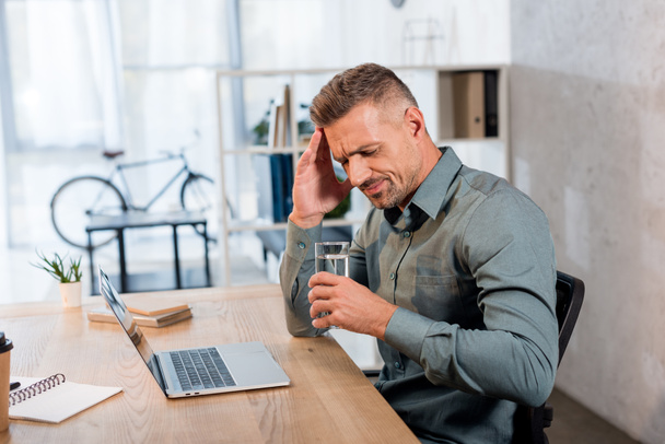exhausted businessman holding glass of water while having headache in office - Foto, Imagem