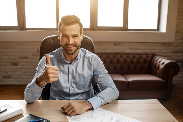 Young handsome cheerful businessman sit on table and pose in his own office. He hold big thumb up and smile. Nice happy guy. Sunlight. - 写真・画像
