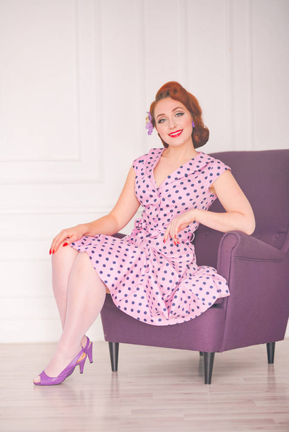 pretty redheaded pin up woman wearing pink polka dot dress and posing with purple armchair on white background - Foto, imagen