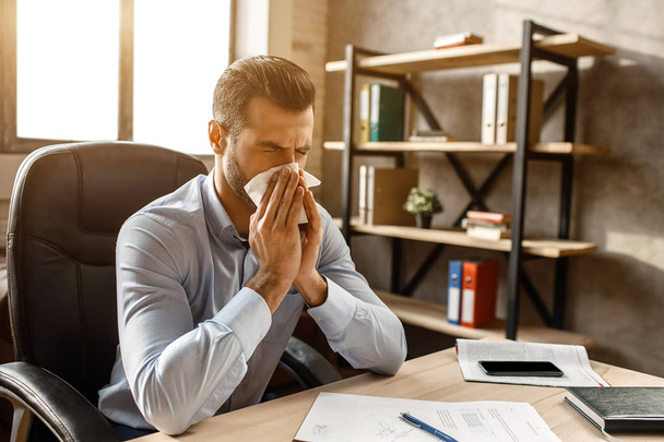 Young handsome businessman sit at table and sneeze in his own office. He cover nose with white napkin. Sick guy suffer. - Photo, Image