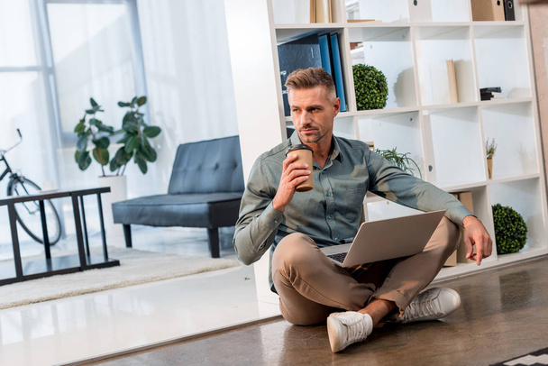 handsome businessman sitting on floor with laptop and coffee to go in office - Foto, immagini