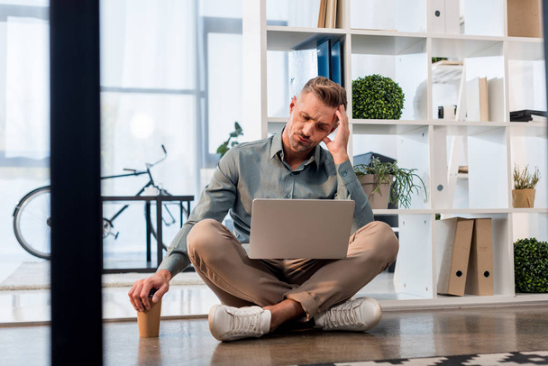 handsome businessman sitting with crossed legs on floor and looking at laptop - Photo, Image