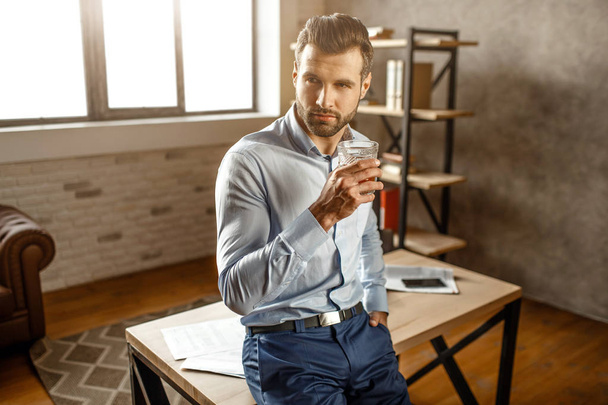 Young handsome businessman stand and hold glass of whiskey in his own office. He look to side and lean to table. Daylight. - Fotoğraf, Görsel