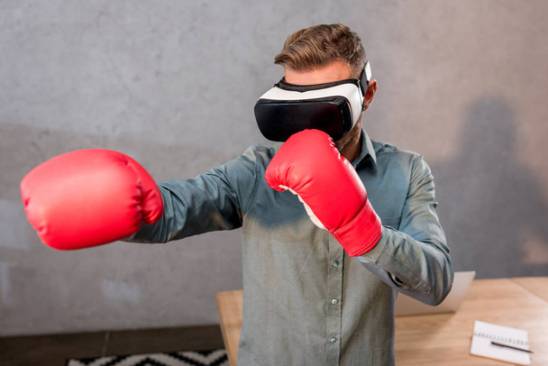 selective focus of businessman wearing virtual reality headset and boxing gloves in office - Фото, изображение