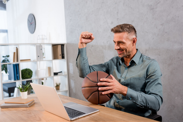 cheerful businessman holding basketball while watching championship on laptop in office - Фото, изображение