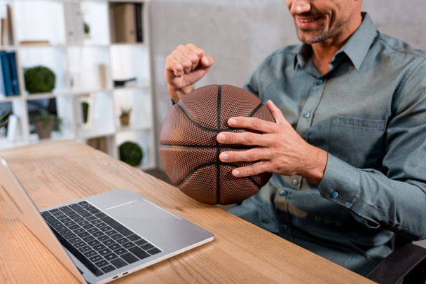 Ausgeschnittene Ansicht eines gut gelaunten Geschäftsmannes, der Basketball hält, während er die Meisterschaft auf seinem Laptop im Büro verfolgt - Foto, Bild