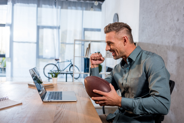 happy businessman watching championship on laptop and holding american football in modern office - Photo, Image