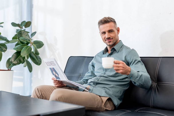 cheerful businessman sitting on sofa while holding business newspaper and cup with drink - Zdjęcie, obraz