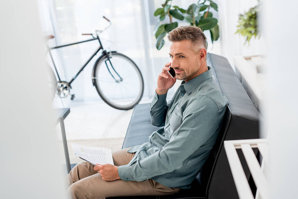 selective focus of businessman holding newspaper and talking on smartphone while sitting on sofa - Valokuva, kuva