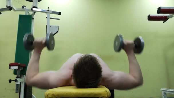 Young athletic man trains pectoral muscles in the gym with dumbbells, engaged in fitness - Video, Çekim