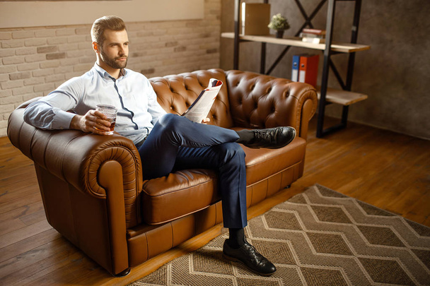 Young handsome buisnessman sitting on sofa and drink whiskey in his own office. He look straight with confidence. Guy hold journal. Attractive small smile. - Photo, image