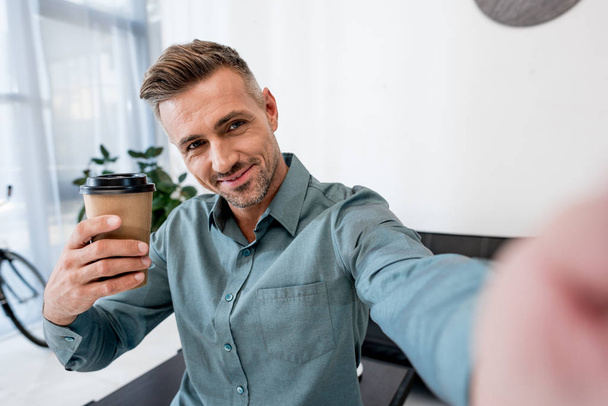 wesoły człowiek mówi selfie trzymając kubek papierowy w nowoczesnym biurze  - Zdjęcie, obraz
